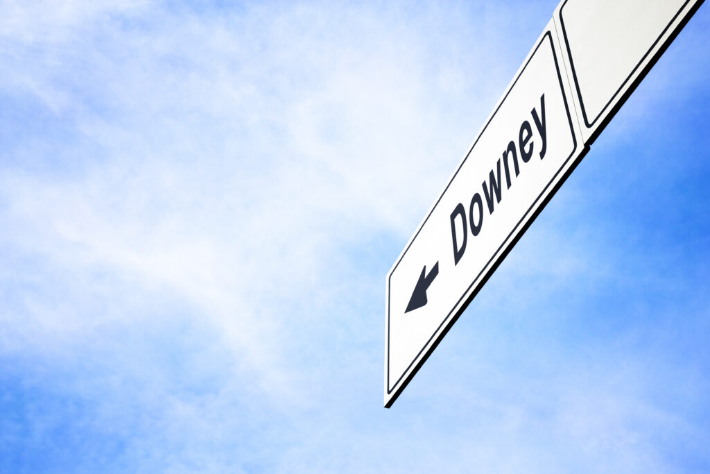 White signboard with an arrow pointing left towards Downey, California, USA, against a hazy blue sky in a concept of travel, navigation and direction. Path included for the signboard
